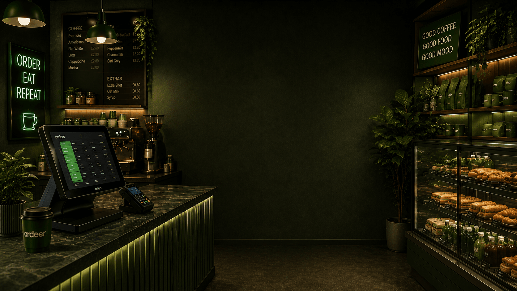 One deep-green coffee shop: the Ordeer till and card terminal on the counter beside the espresso machine and menu board, with the bakery cabinet and shelves of beans on the right.
